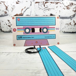 May include: A white cassette tape with blue and purple stripes. The tape is labeled "UR POSITION NORMAL TYPE I" and "HIGH BIAS 70&mu;S EQ". The tape is 90 minutes long. The tape is attached to three blue and white striped ribbons.