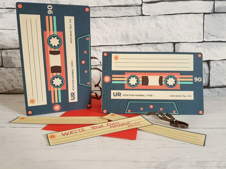 May include: Two blue and orange cassette tape cards with the text "UR POSITION NORMAL TYPE I HIGH BIAS 70US EQ" and "90". The cards are on a brick wall background. There are also several blank cards with the text "WRITE YOUR OWN MESSAGE HERE" on a wooden surface.