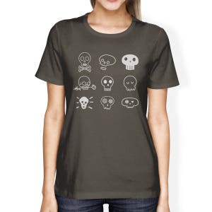 May include: A dark gray t-shirt with a white design of nine different skull illustrations. The skulls are all different shapes and sizes, and some have flowers or other details.