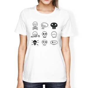 May include: White t-shirt with a black and white design of various skull illustrations. The skulls have different features, including flowers, crossbones, and eyes.