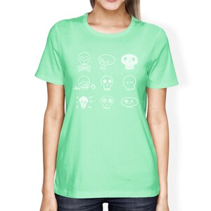 May include: A light green t-shirt with a white design of various skull illustrations. The skulls have different facial expressions and details, including crossbones, flowers, and hearts.
