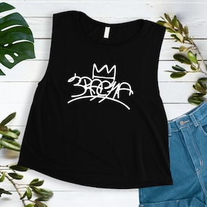 Può includere: Canotta nera senza maniche con una grafica bianca di una corona e la parola "3REYA" in stile graffiti.