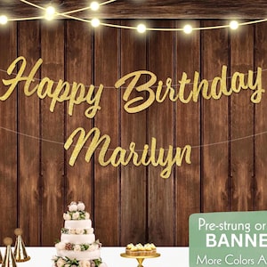 May include: A gold glitter "Happy Birthday Marilyn" banner strung across a wooden backdrop, with string lights overhead. A multi-tiered cake and other party decorations are in the foreground. The banner is a festive decoration for a birthday celebration.