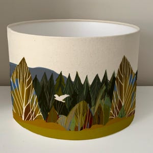 May include: A white lampshade with a forest scene printed on it. The scene features trees, a bird flying, and a yellow, green, and brown color scheme.