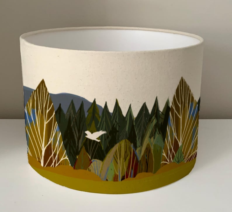 May include: A white lampshade with a forest scene printed on it. The scene features trees, a bird flying, and a yellow, green, and brown color scheme.