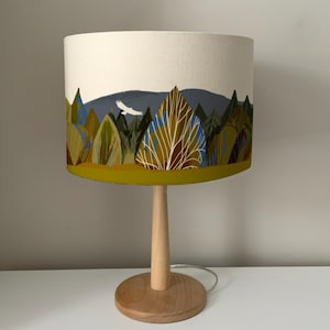 May include: A table lamp with a white drum shade featuring a forest scene with green, blue, and yellow colors. The lamp base is made of natural wood.