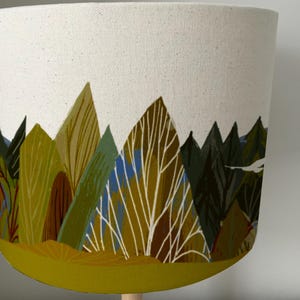 May include: A white lampshade with a forest scene printed on it. The scene features green, brown, and yellow trees and a white sky. The bottom of the lampshade is a solid yellow color.