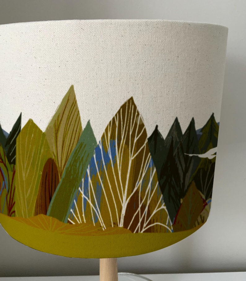 May include: A white lampshade with a forest scene printed on it. The scene features green, brown, and yellow trees and a white sky. The bottom of the lampshade is a solid yellow color.