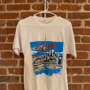 May include: White t-shirt with a graphic print of the Toronto skyline, including the CN Tower, and the text "Toronto" and "Canada".