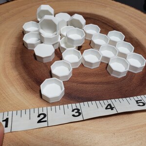 May include: A group of 20 white hexagonal plastic containers, some stacked on top of each other, on a wooden surface. A measuring tape is partially visible in the foreground.