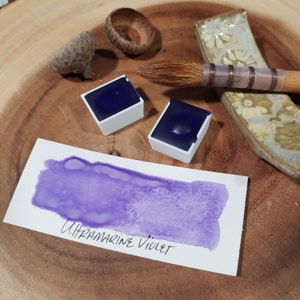 May include: Two small pans of ultramarine violet watercolor paint, with a sample of the color painted on a white card. The card reads "Ultramarine Violet".