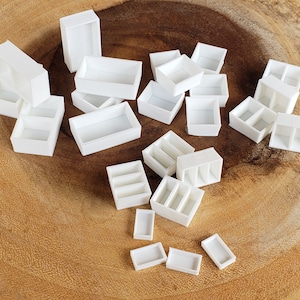 May include: A collection of white plastic paint palettes of various sizes and configurations. Some palettes have multiple wells, while others have a single large well. The palettes are arranged on a wooden surface.