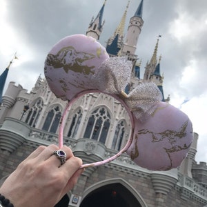 May include: Pink Minnie Mouse ears with a gold glitter design and a sparkly silver bow. The ears are held up in front of Cinderella's Castle at Walt Disney World.