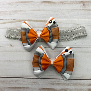 May include: Two orange, white, and gray striped ribbon bows with black buttons. The bows are attached to a white lace headband.