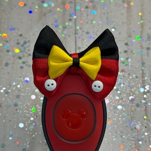 Pode incluir: Um acessório de pulseira vermelho e preto com um laço de tecido decorativo com um centro amarelo e dois pequenos botões brancos. A pulseira tem uma peça central circular vermelha gravada com a silhueta de uma cabeça de rato.