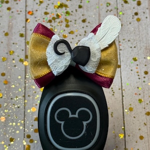 May include: A decorative bow accessory featuring gold and maroon fabric, white lace trim, a black hook-shaped charm, and a white feather-shaped accent, attached to a black wearable device with a circular Mickey Mouse icon on the front face.