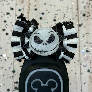 May include: A black and white striped bow featuring a Jack Skellington face. The bow is attached to a black MagicBand with a Mickey Mouse icon.