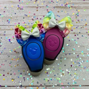 May include: Two colorful Disney park MagicBands with rainbow patterned Minnie Mouse ears attached. The MagicBands are blue and purple with Mickey Mouse ears on the front. The background is a light wood surface with colorful confetti.