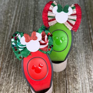 May include: Two Disney MagicBands, one red and one green, each adorned with a festive bow. The red band features a Mickey Mouse icon, while the green band has a Mickey Mouse icon. The bows are red, green, and white, with glitter accents.