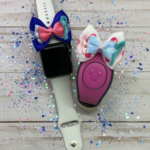 May include: Two Disney-themed accessories for a smartwatch and a MagicBand. The smartwatch band is white with a blue and pink bow with a blue number "1" on it. The MagicBand is pink with a white and blue bow with a blue number "1" on it. Both bows have a white lace overlay.