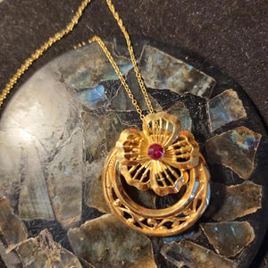May include: A gold-colored pendant necklace featuring a floral design with a central red gemstone. The pendant is suspended from a delicate gold chain. The necklace is displayed on a dark, textured surface.