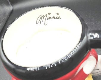 May include: A ceramic mug shaped like Minnie Mouse, featuring a black rim, a red body with white polka dots, and a yellow base. The interior is white with the word Minnie printed in black script, accompanied by two small hearts above the text.