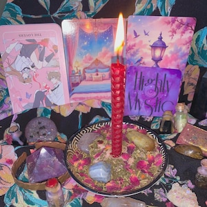 May include: A red textured candle burns in a bowl filled with dried rose petals and stones. Behind it are three illustrated cards, one showing a bedroom, one with the text Highly Mystic, and one titled The Lovers, alongside various crystals.