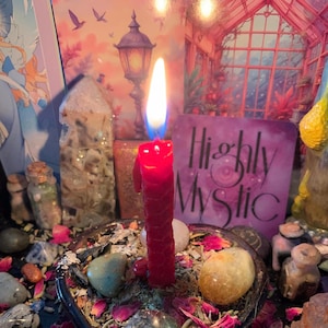 May include: A lit red candle with a long, slender shape, surrounded by crystals, stones, and dried petals. The candle's flame is bright, with a blue base. A card with the words "Highly Mystic" is visible.