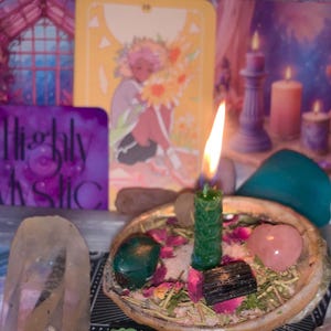 Puede incluir: Una escena espiritual con una vela verde encendida, un cuarzo rosa en forma de corazón y un cristal pulido. Una carta de tarot con una ilustración floral y las palabras "Highly Mystic" también son visibles, junto con otros elementos decorativos.