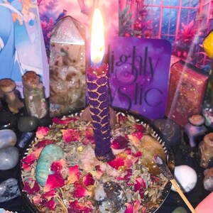 May include: A lit purple candle with a gold honeycomb pattern sits in a black bowl filled with dried herbs, rose petals, and crystals. The candle flame glows brightly. A purple card with the text "Highly Mystic" is in the background.