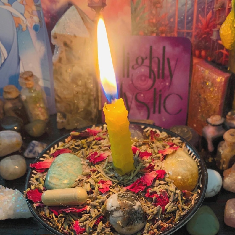 Puede incluir: Una vela amarilla con una llama encendida se encuentra en un cuenco negro lleno de lavanda seca, p&eacute;talos de rosa rojos, una piedra verde claro, una piedra amarilla, una piedra moteada en blanco y negro y una c&aacute;psula de color beige.