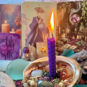 May include: A purple honeycomb-textured beeswax candle with a lit flame, standing upright inside a large iridescent abalone shell. The shell contains a mixture of dried flower petals, small polished gemstones, and smooth river stones.