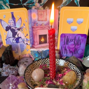 May include: A red honeycomb-textured candle burns in a black and white patterned bowl filled with dried flower petals and smooth stones. Behind the candle are three tarot-style cards, one featuring a fairy, one a fireplace, and one with the text Highly Mystic.