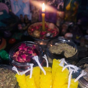 Puede incluir: Un grupo de velas de cera de abeja de color amarillo brillante y texturizadas con mechas de algod&oacute;n blanco, colocadas frente a varios frascos de vidrio con hierbas secas y p&eacute;talos de flores, con una vela amarilla encendida al fondo.