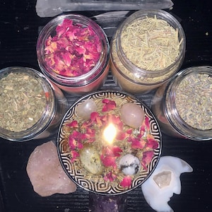 Puede incluir: Una vela encendida en un cuenco estampado cubierto con p&eacute;talos de rosa secos y cristales, rodeada por tres frascos de vidrio transparente con hierbas y flores secas, un trozo grande de cuarzo rosa en bruto y una piedra tallada en forma de luna.