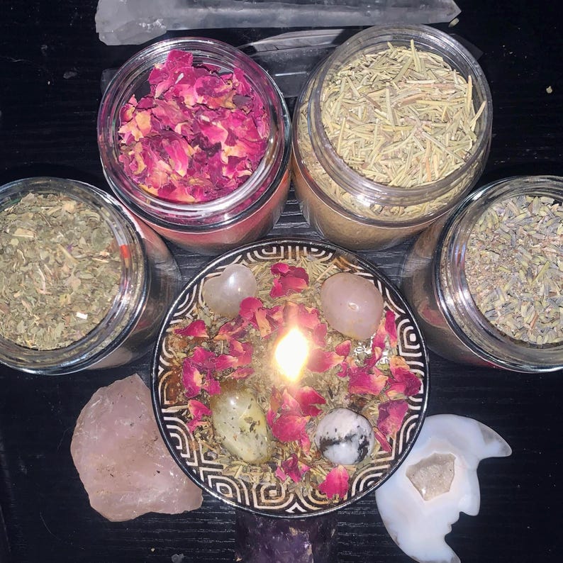 Puede incluir: Una vela encendida en un cuenco estampado cubierto con p&eacute;talos de rosa secos y cristales, rodeada por tres frascos de vidrio transparente con hierbas y flores secas, un trozo grande de cuarzo rosa en bruto y una piedra tallada en forma de luna.