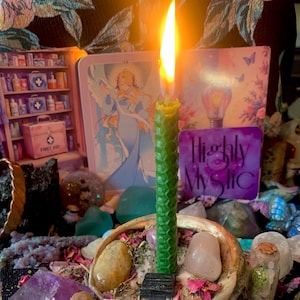 Puede incluir: Una vela de cera de abeja alta, verde y texturizada con una llama encendida, colocada en un cuenco de concha lleno de varias piedras pulidas, cristales y p&eacute;talos de flores secas, frente a varias tarjetas ilustradas coloridas.