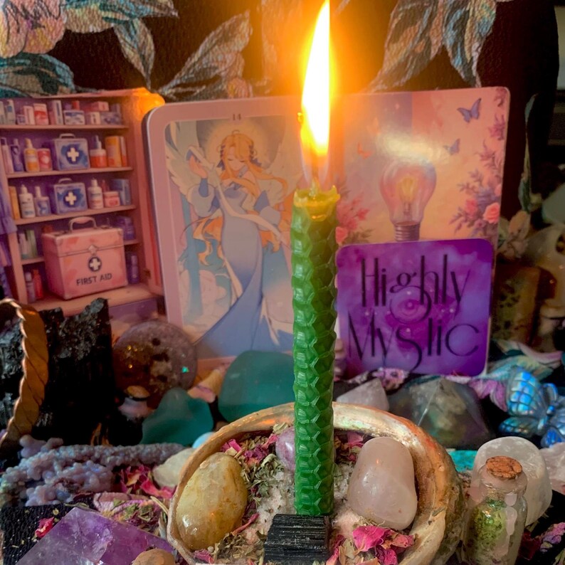 Puede incluir: Una vela de cera de abeja alta, verde y texturizada con una llama encendida, colocada en un cuenco de concha lleno de varias piedras pulidas, cristales y p&eacute;talos de flores secas, frente a varias tarjetas ilustradas coloridas.