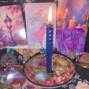 Può includere: Una candela blu accesa con una superficie strutturata si trova in una ciotola con cristalli, pietre e petali di rosa secchi. Carte dei tarocchi e altre candele sono sullo sfondo. La carta viola dice "Highly Mystic".