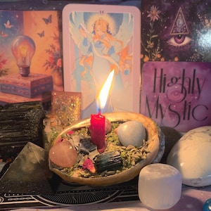 Puede incluir: Una vela roja encendida se encuentra en una concha decorativa llena de cristales y hierbas. Las cartas del tarot y otros objetos espirituales están dispuestos alrededor de la concha. Las palabras "Highly Mystic" son visibles en una carta.