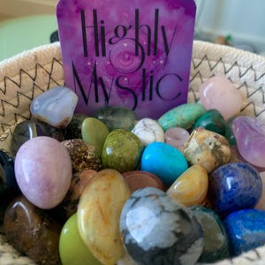 Puede incluir: Una cesta tejida llena de una variedad de piedras pulidas y cristales multicolores. Detr&aacute;s de las piedras hay una tarjeta rectangular morada con el texto Highly Mystic impreso en negro, que presenta un gr&aacute;fico de un ojo central.