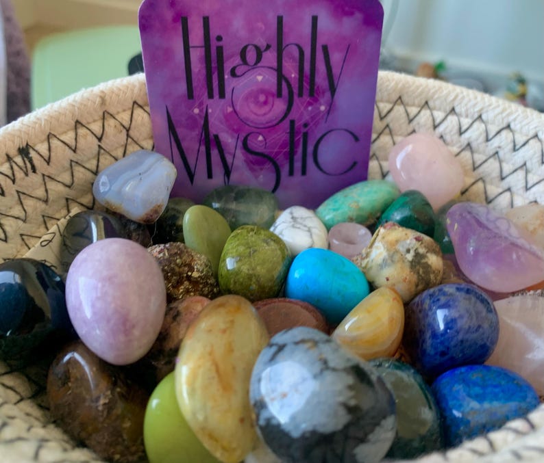 Puede incluir: Una cesta tejida llena de una variedad de piedras pulidas y cristales multicolores. Detr&aacute;s de las piedras hay una tarjeta rectangular morada con el texto Highly Mystic impreso en negro, que presenta un gr&aacute;fico de un ojo central.