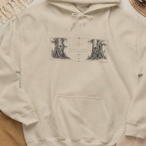 Pode incluir: Uma camisola com capuz de cor creme com um gráfico em estilo de tinta preta no peito. O design retrata dois rostos de árvores antigas dentro de uma moldura retangular, rodeados por pequenas estrelas e símbolos celestiais, com texto no centro.