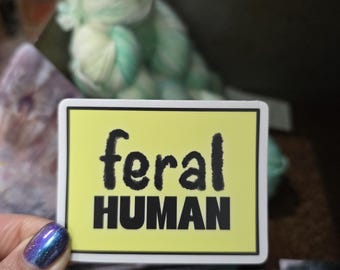 Puede incluir: Una pegatina rectangular con fondo amarillo pálido y un borde negro grueso. La palabra feral está impresa en una fuente negra de estilo manuscrito sobre la palabra HUMAN, escrita en letras mayúsculas negras, negritas y sin remates.