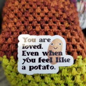 May include: A crocheted item in shades of purple, orange, and green. A white sticker with the text "You are loved. Even when you feel like a potato" and a smiling potato illustration is attached to the item.