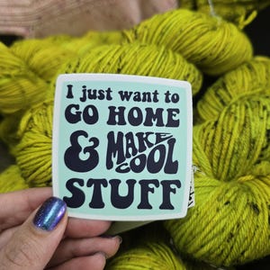 May include: A sticker with the text "I just want to go home & make cool stuff" in a retro font. The sticker is held in front of a pile of bright yellow-green yarn. The yarn is in a skein.