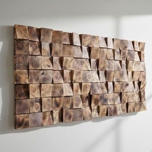 Puede incluir: Una pieza de arte mural rectangular hecha de numerosos bloques de madera cuadrados y pequeños. Los bloques están dispuestos en un patrón de mosaico, mostrando la veta natural de la madera y varios tonos de marrón. La obra está montada en una pared blanca.