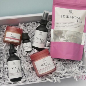 May include: A white box filled with shredded paper, containing skincare products. The products include a jar of "icaboo" balm, a bottle of "scalp oil", a bottle of "cooling spray", a jar of "body lotion", a small bottle of "hormone blend", and a pink bag of "hormone tea".