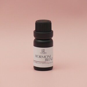 Pu&ograve; includere: Piccola bottiglia di olio essenziale nero con tappo nero ed etichetta bianca. L'etichetta recita "Hormone Blend" con testo aggiuntivo sottostante. La bottiglia &egrave; posta su uno sfondo rosa tenue. Il prodotto &egrave; probabilmente per aromaterapia o benessere.
