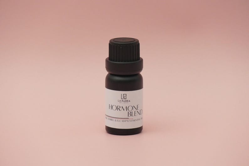 Pu&ograve; includere: Piccola bottiglia di olio essenziale nero con tappo nero ed etichetta bianca. L'etichetta recita "Hormone Blend" con testo aggiuntivo sottostante. La bottiglia &egrave; posta su uno sfondo rosa tenue. Il prodotto &egrave; probabilmente per aromaterapia o benessere.